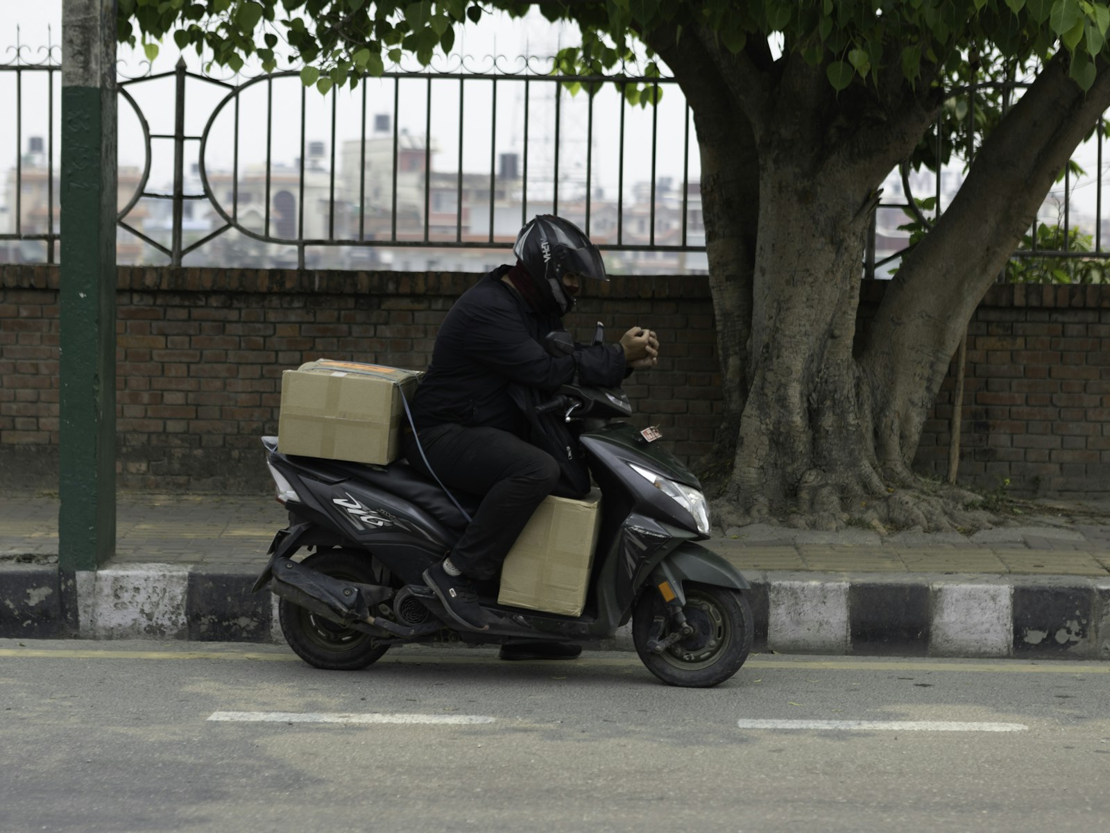 Delivery rider in a helmet on a scooter carrying cardboard parcels along a tree-lined South Asian city street