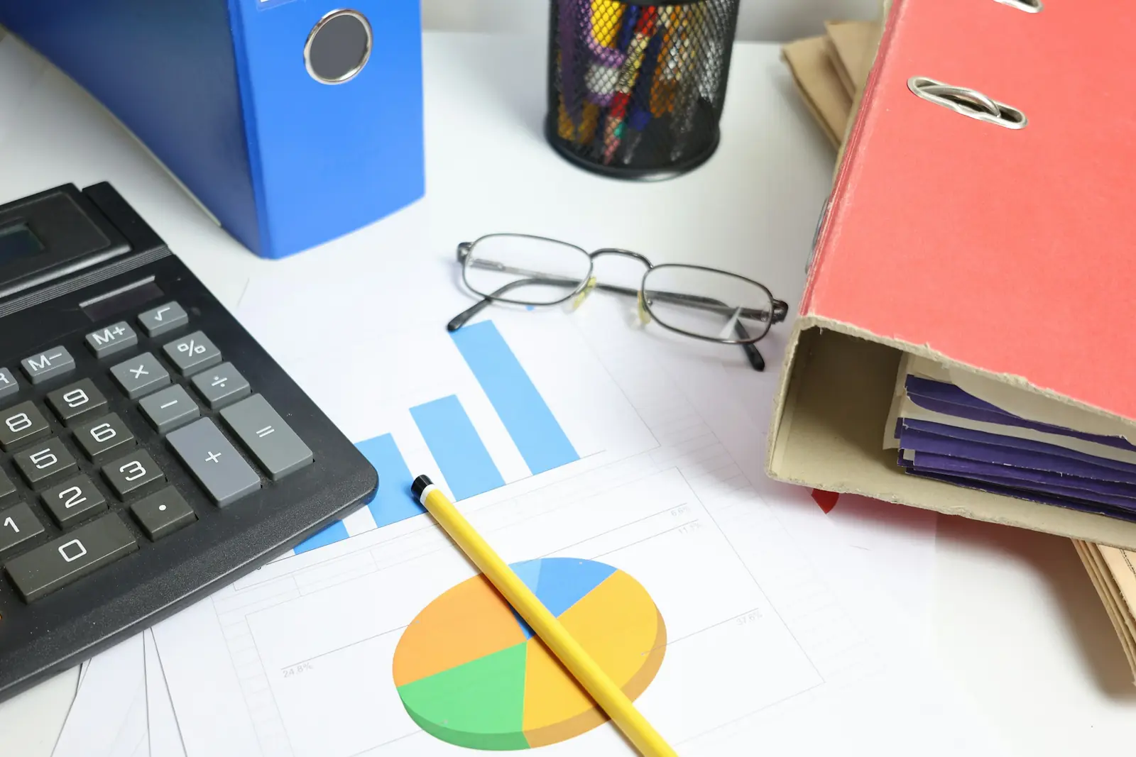 Desk calculator, printed bar and pie charts, reading glasses and bulging ring binders on an accountant's desk