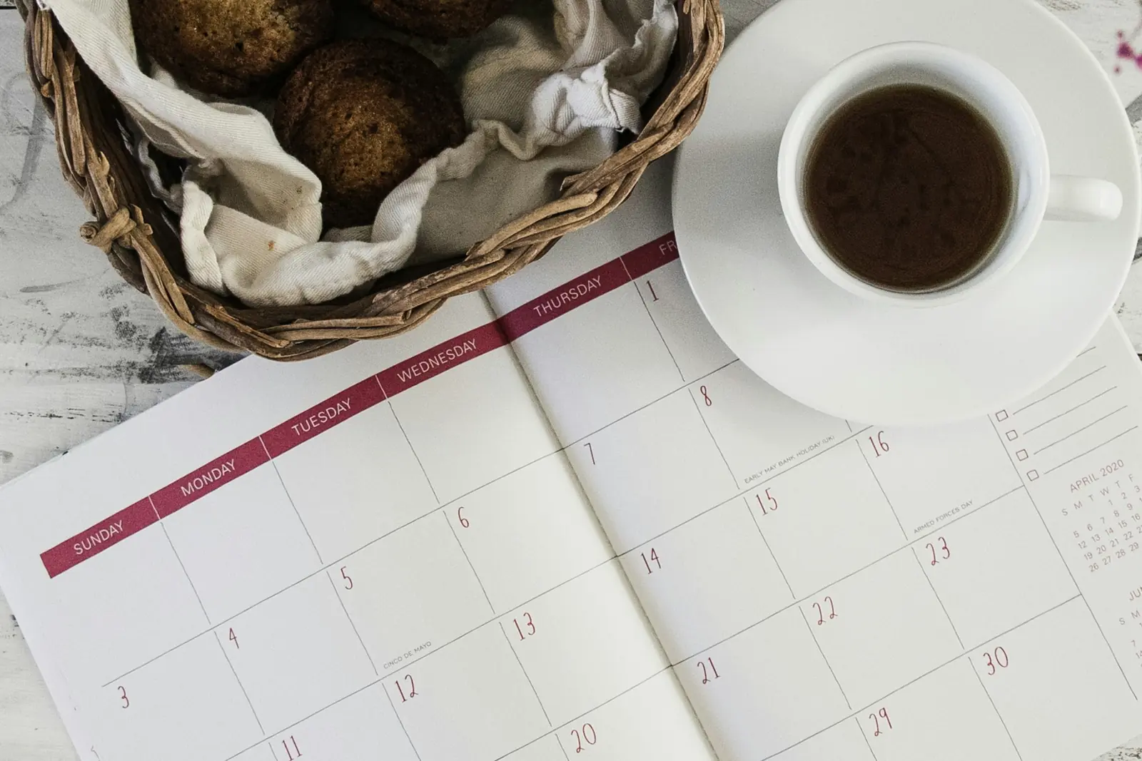 Open monthly planner with red weekday headers beside a cup of black tea and a basket of muffins, seen from above