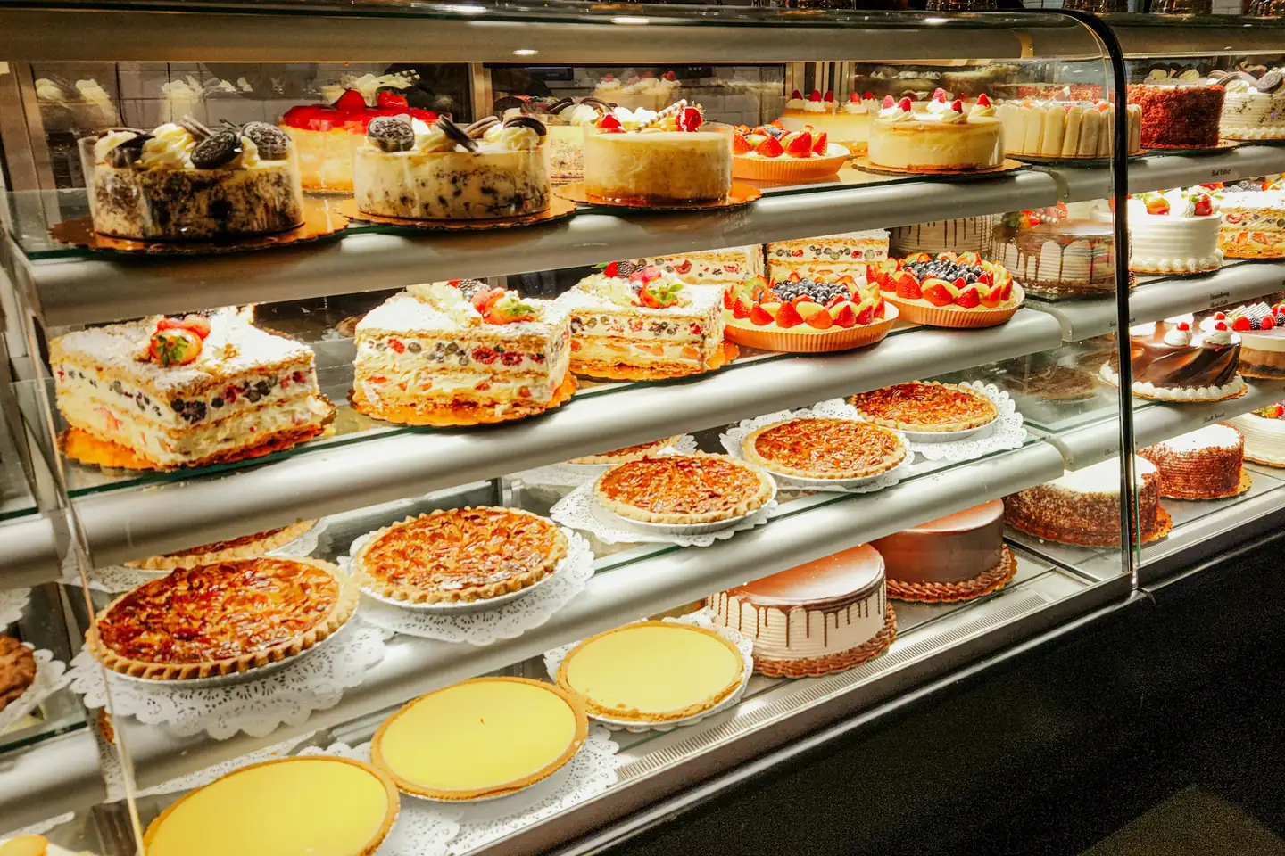 Glass bakery display case of layered cream cakes, fruit tarts and baked pies arranged on paper doilies