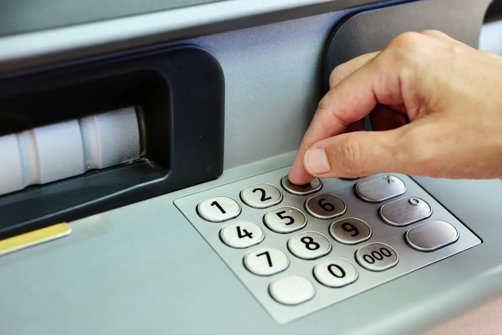 Close-up of a hand entering a PIN on the steel number keypad of a cash machine beside the cash slot