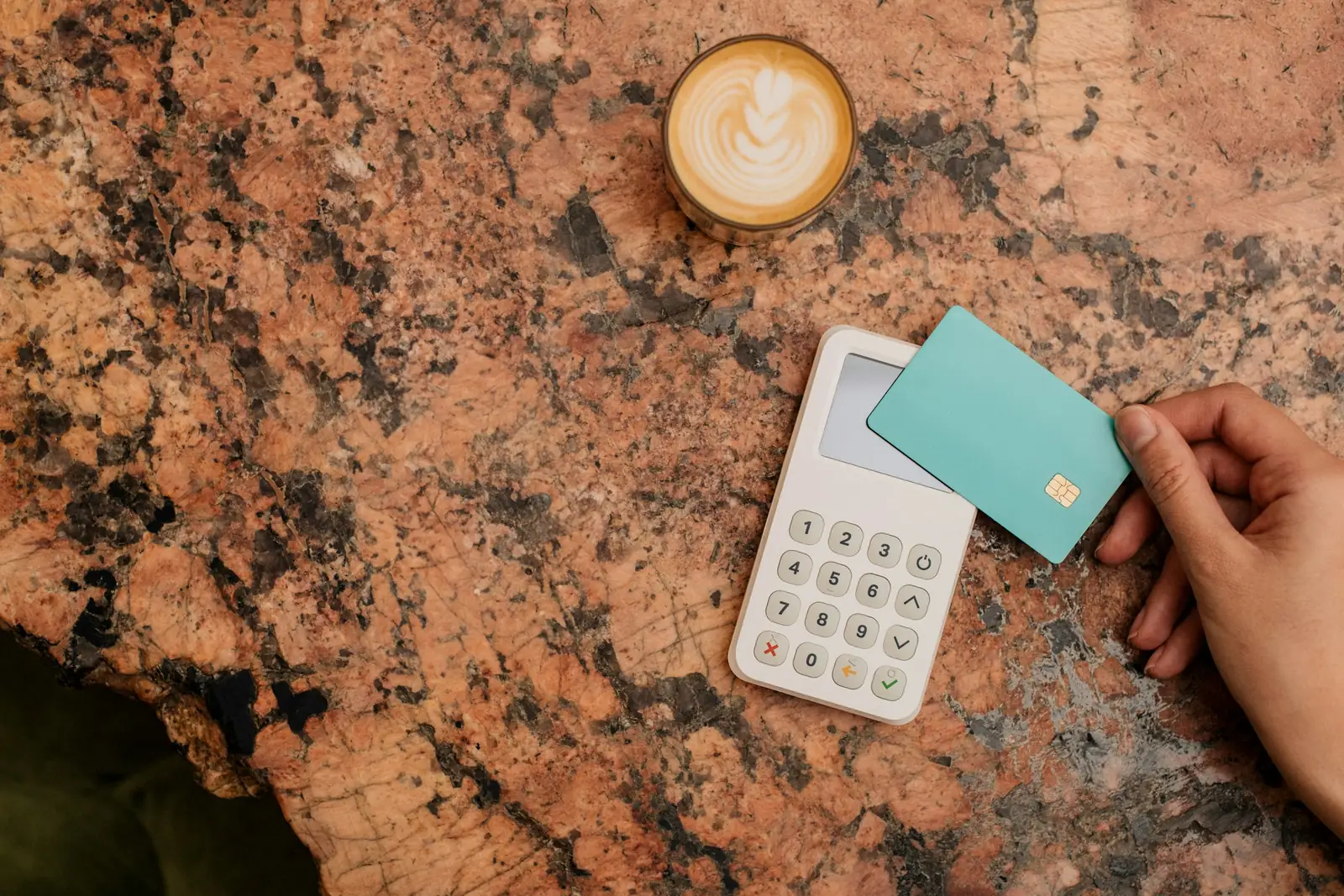 Hand tapping a plain teal chip card on a white keypad card reader beside a cortado on a granite café table