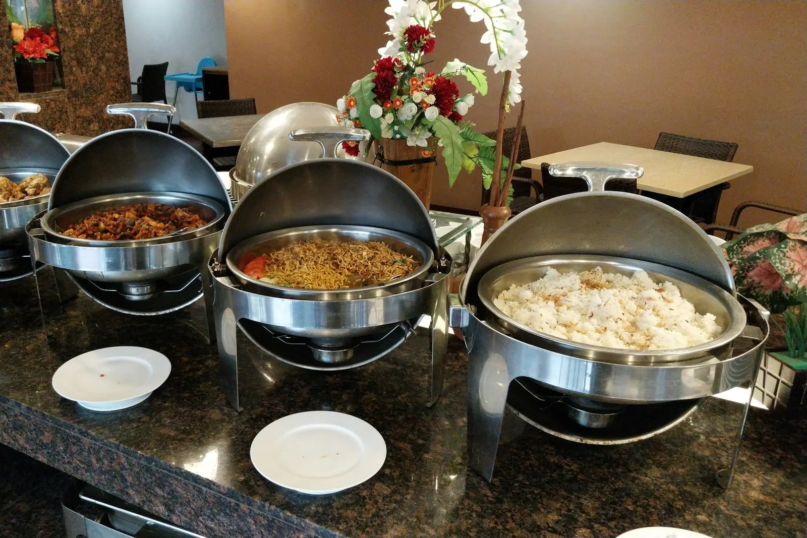 Row of steel chafing dishes with open lids holding noodles, rice and stir-fried dishes on a catering buffet