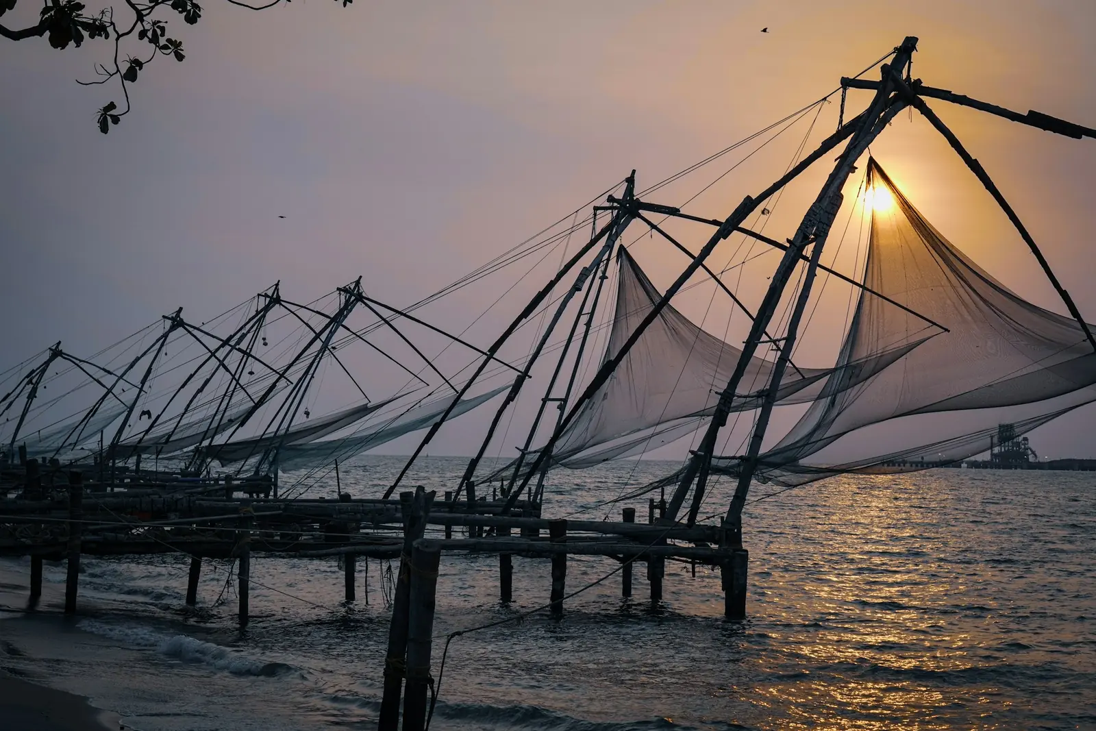 Row of Chinese fishing nets on timber platforms at Fort Kochi, silhouetted against the setting sun over the sea