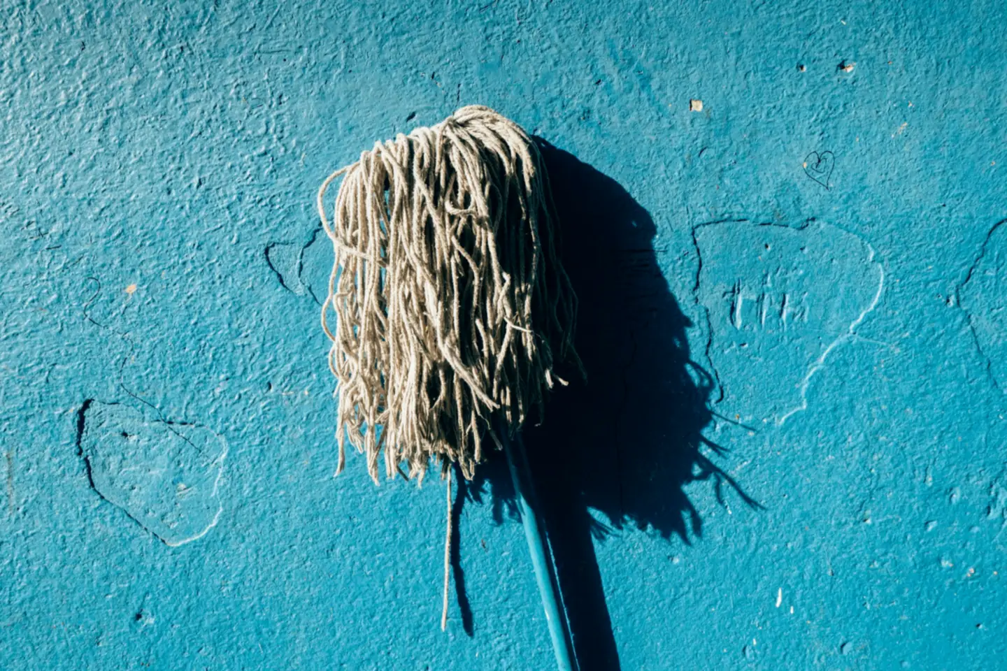 Cotton string mop leaning against a sunlit blue plaster wall, casting a long sharp shadow beside it