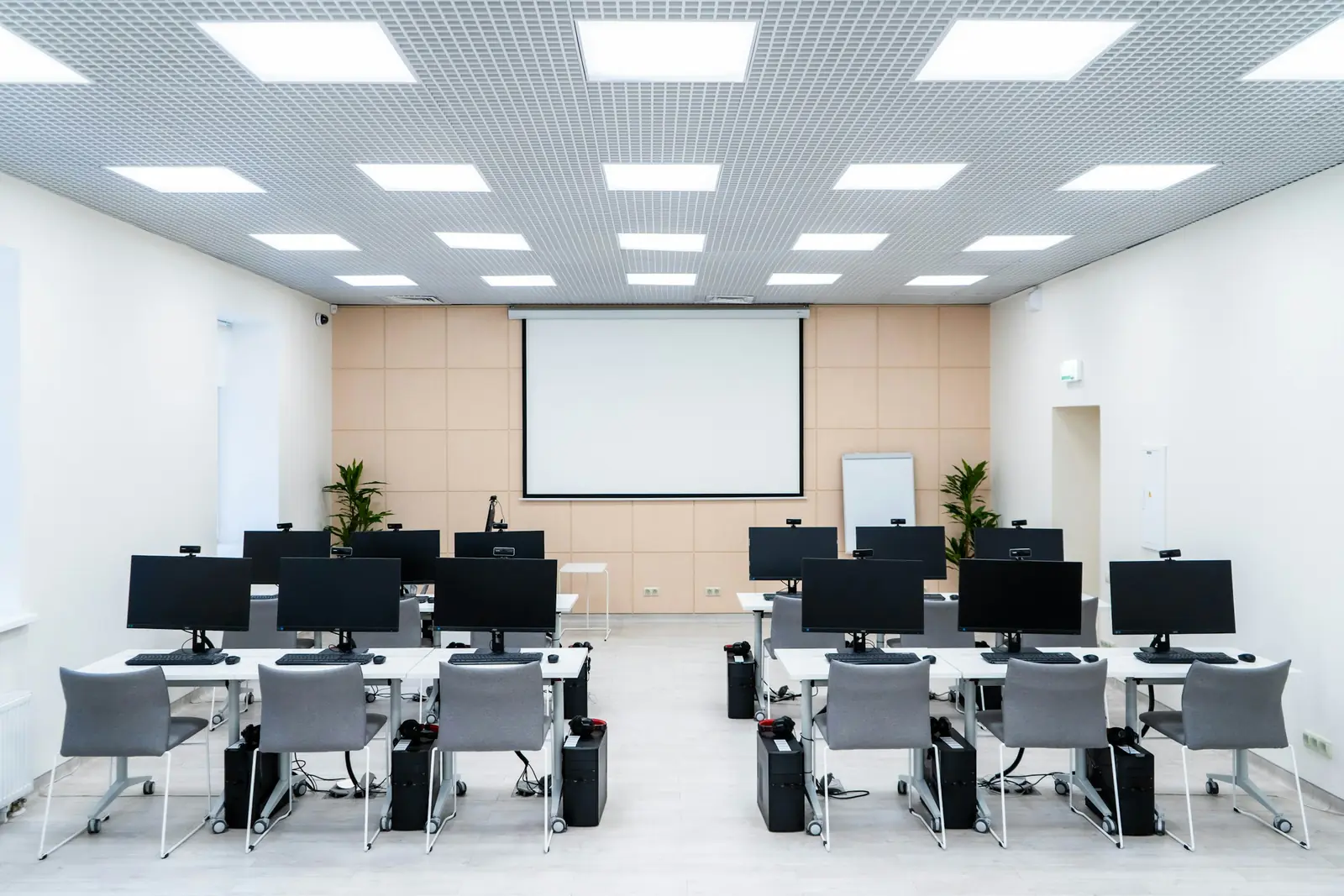 Empty computer lab classroom with rows of monitors, webcams and grey chairs facing a projector screen