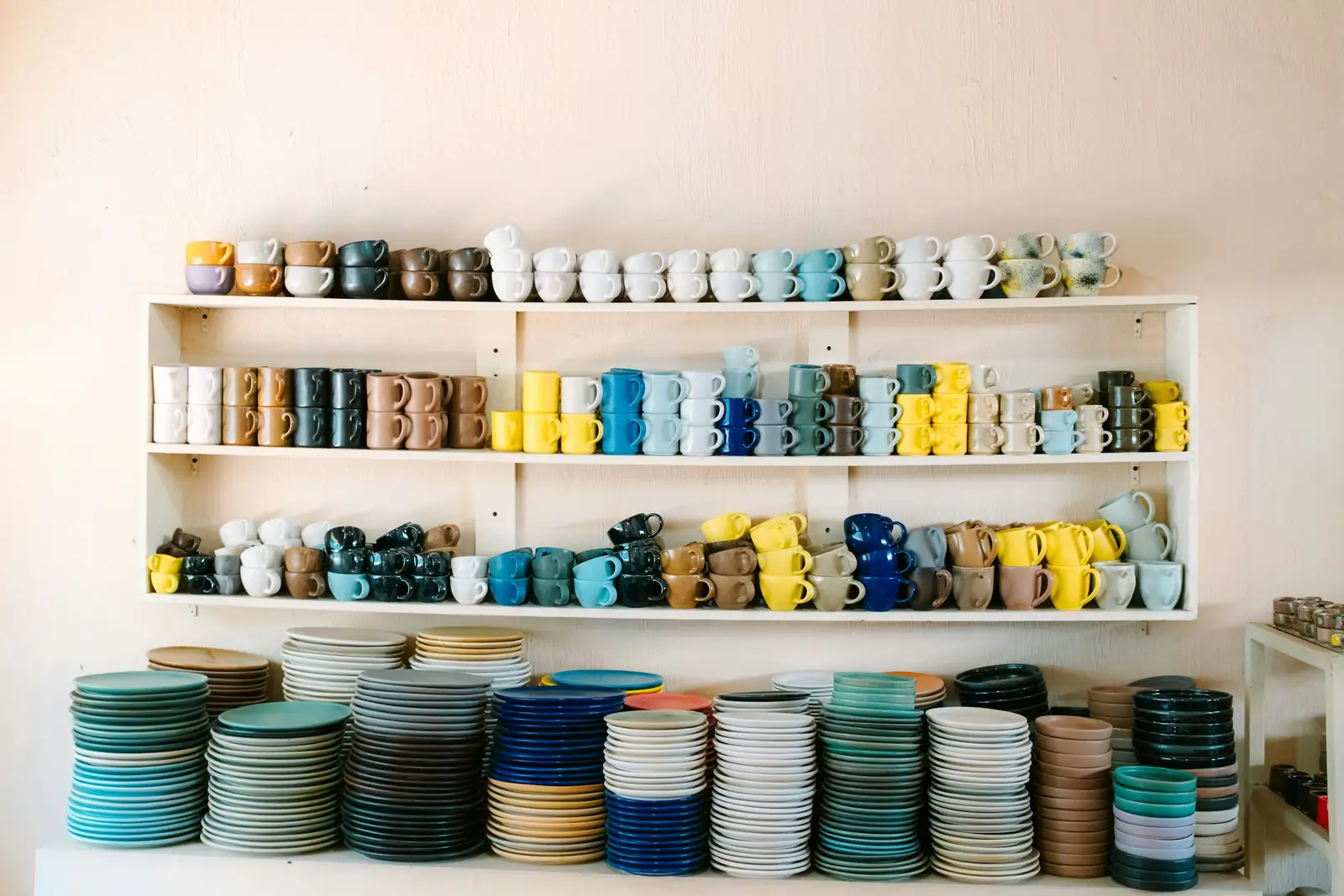 Wall shelves of colourful ceramic mugs above tall stacks of plates in a homeware store