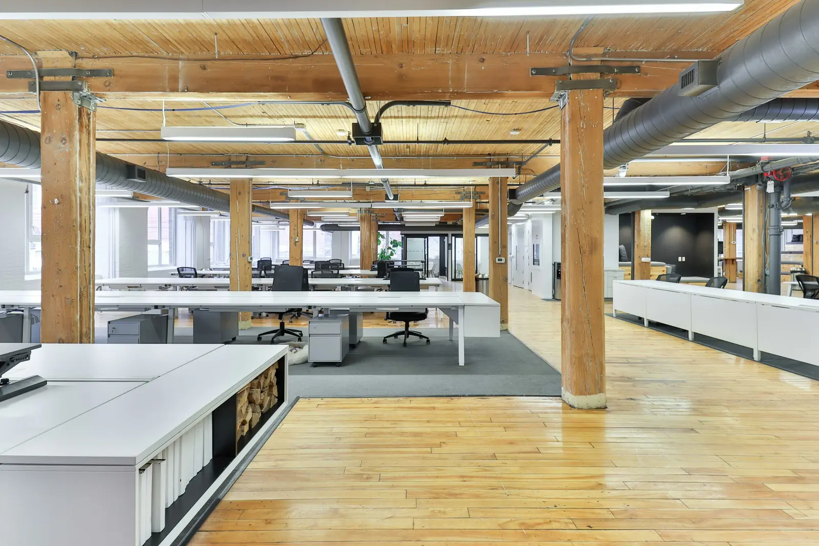 Empty open-plan office in a converted warehouse, with rows of white desks between timber columns and ducting