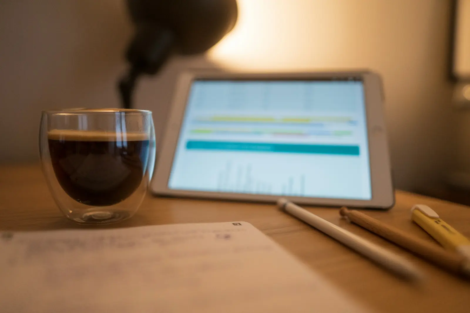 Tablet showing a lesson chart beside a glass of coffee, a stylus, a pencil and notes on a study desk at night