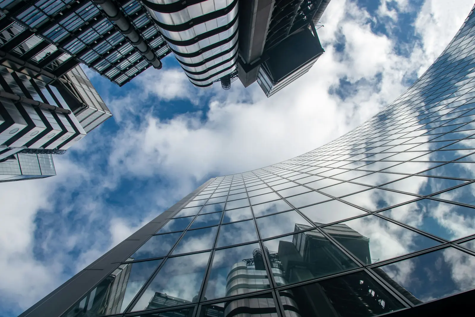 Looking up at curved glass office towers that reflect a blue sky full of white clouds in a financial district