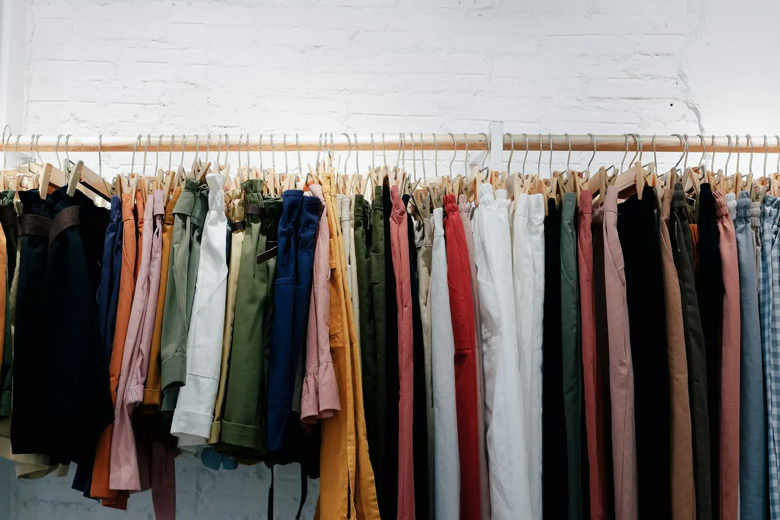 Rail of colourful trousers and shirts on wooden hangers against a white brick wall in a clothing store