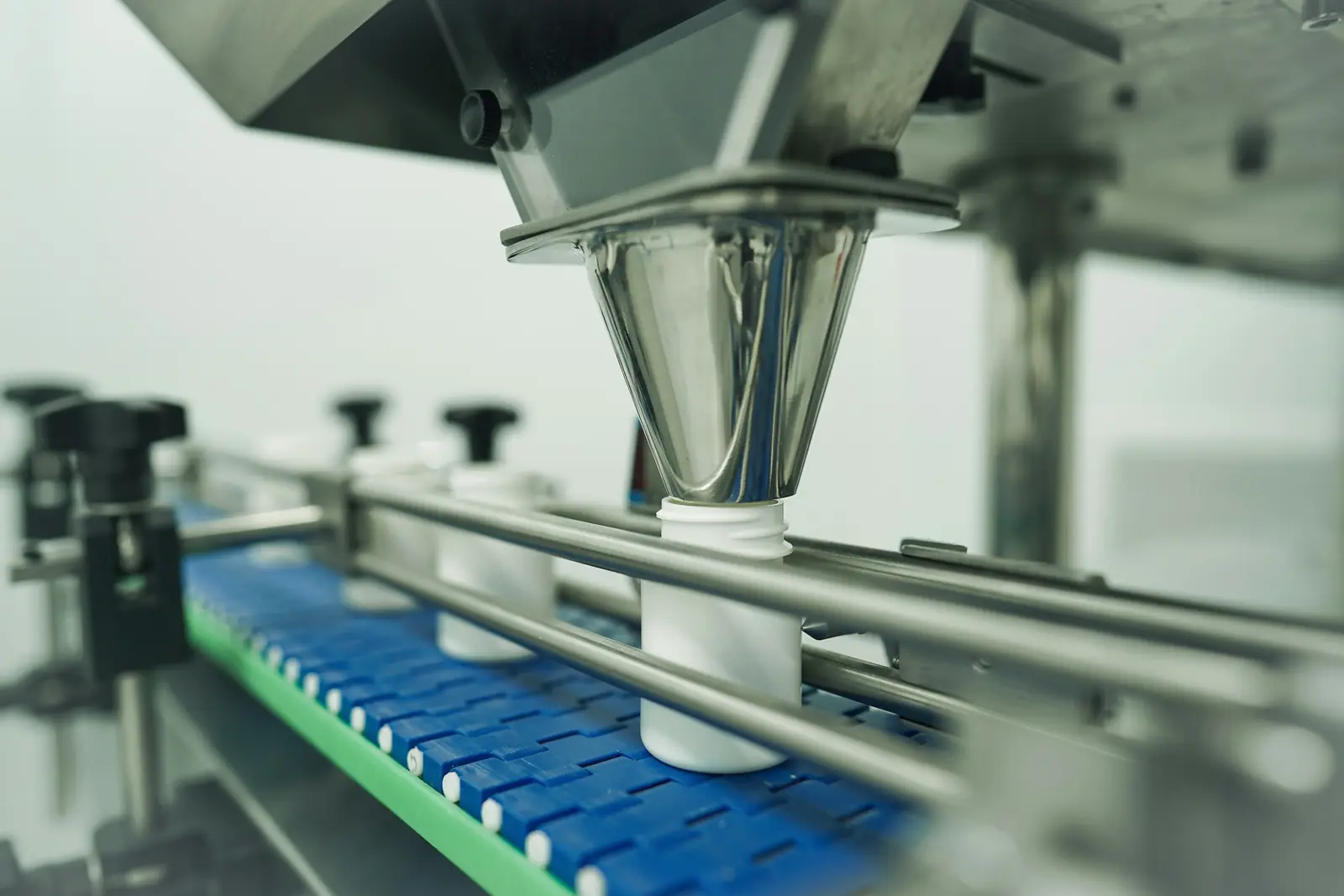 Stainless steel filling funnel dispensing into white plastic containers on a blue conveyor in a packaging plant
