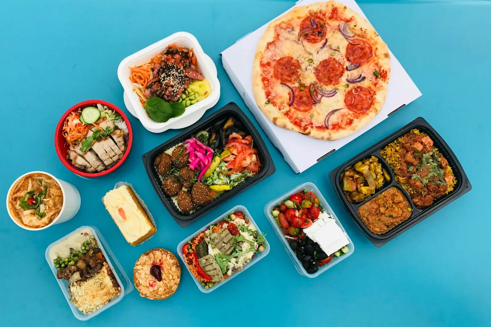 Takeaway containers of salads, curry, rice bowls and a boxed pizza laid out on a blue surface, seen from above