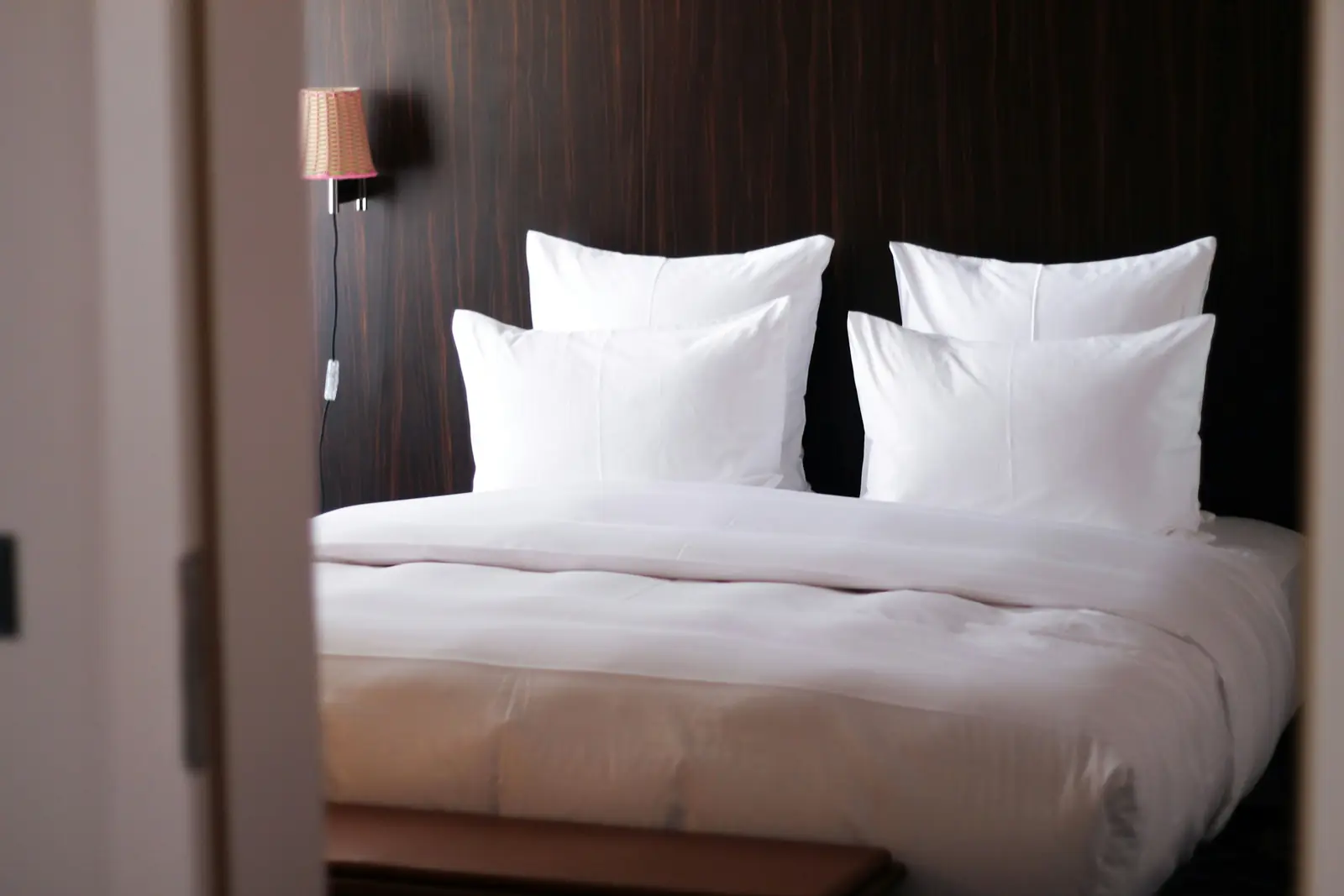 Freshly made hotel bed with crisp white linen and four pillows against a dark wood headboard wall