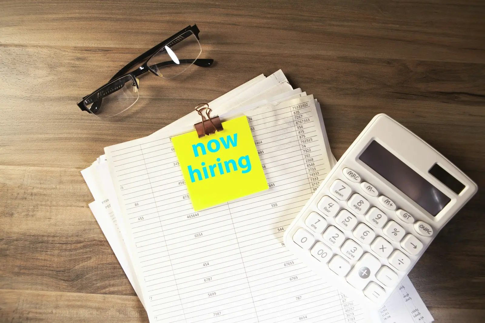 Yellow sticky note reading now hiring clipped to printed spreadsheets beside a calculator and reading glasses