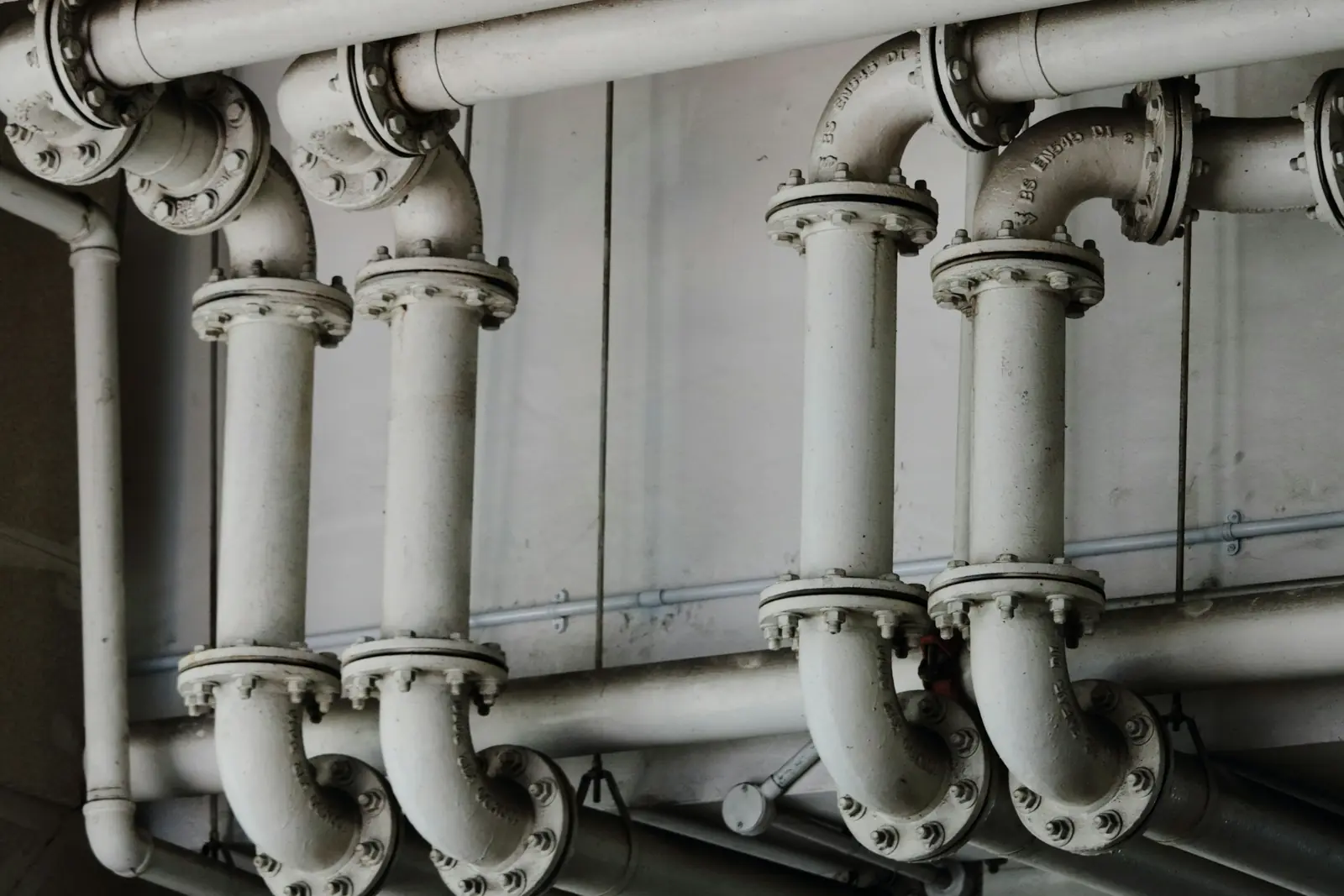 Grey cast-iron pipework with bolted flanges and U-bends running along the wall of an industrial plant room