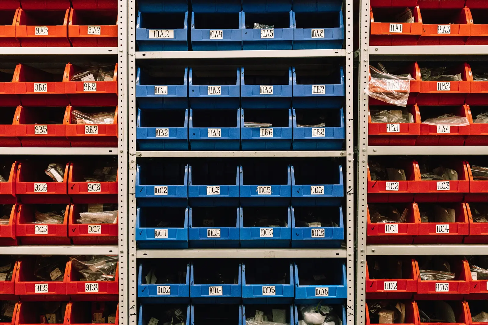 Stockroom shelving of red and blue parts bins, each tagged with a handwritten bin location code such as 10.B.3