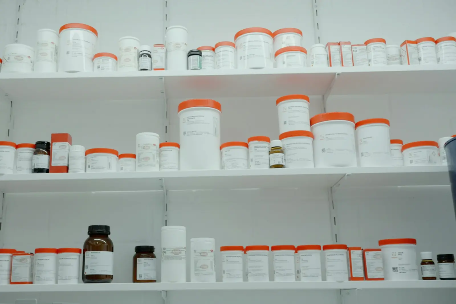 White pharmacy shelves stocked with medicine tubs and bottles with orange lids and amber glass jars