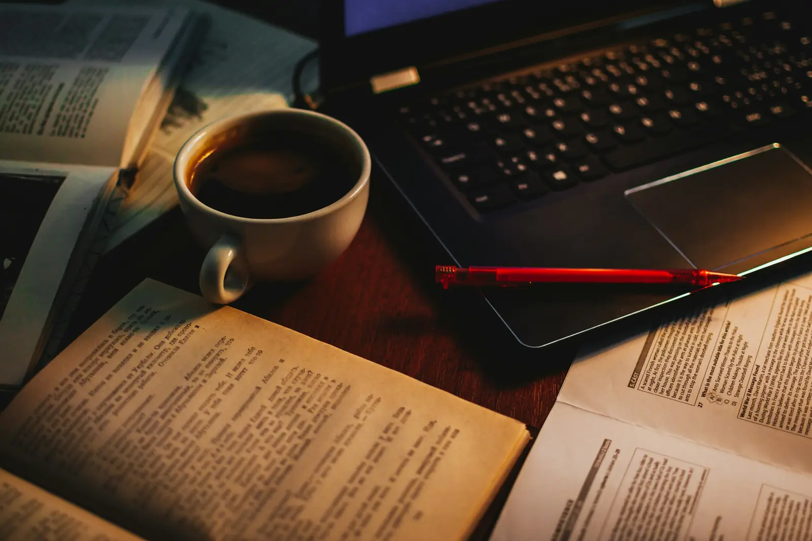 Open textbooks, printed exam papers, a red pen and a cup of coffee beside a laptop on a desk at dusk
