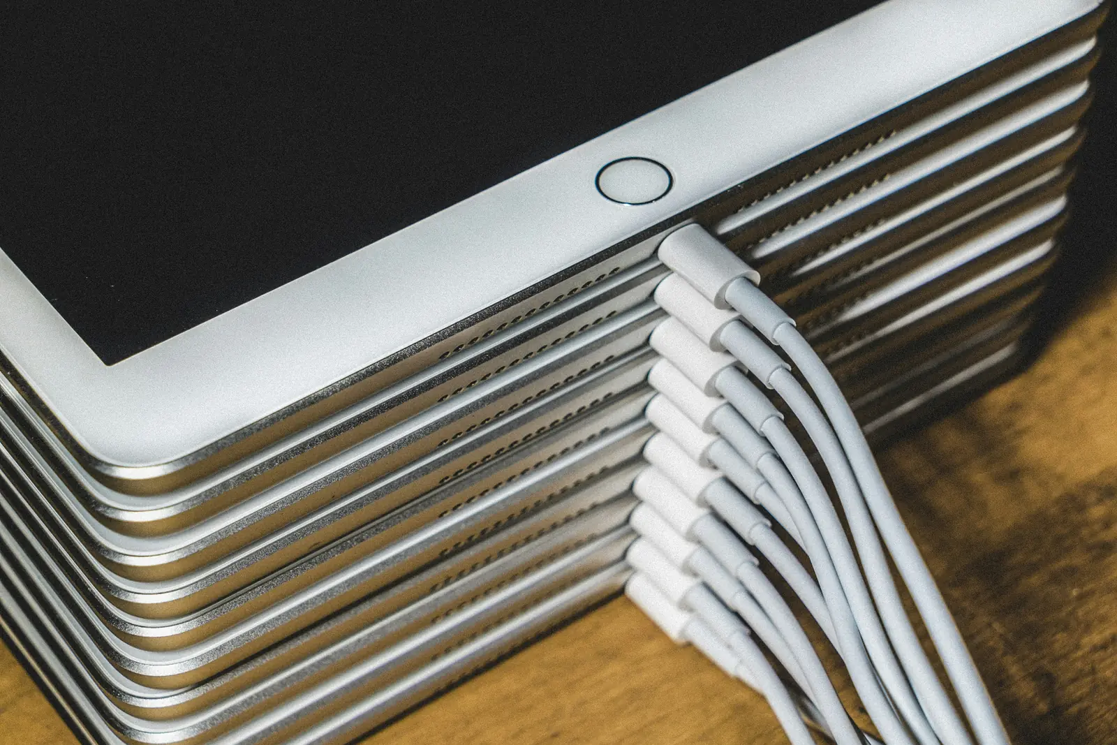 Stack of ten school tablets on a wooden desk, each plugged into its own white charging cable