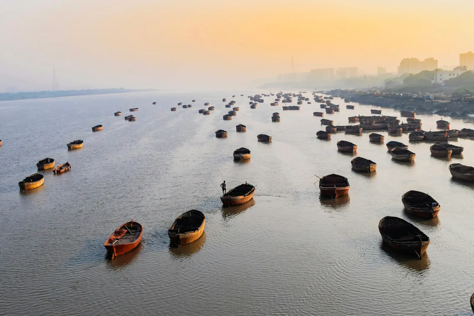 Wooden boats moored across the Tapi River in Surat at sunrise, with the hazy city skyline along the right bank