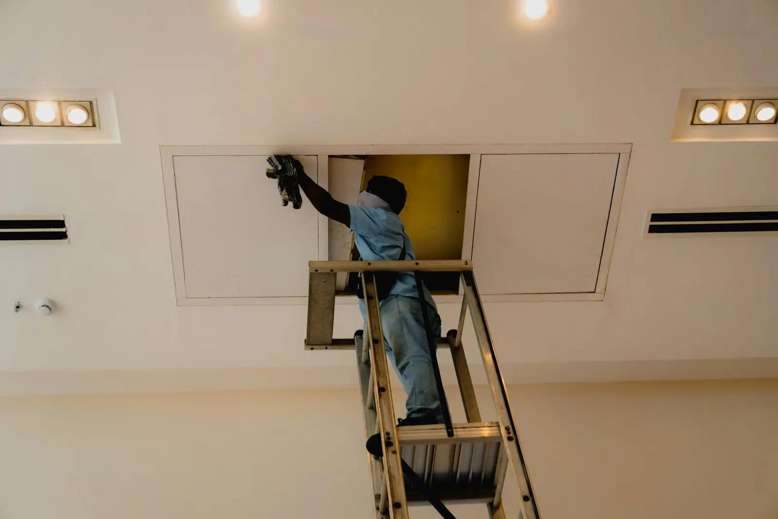 Service technician on an aluminium ladder cleaning inside an open ceiling access panel between recessed lights