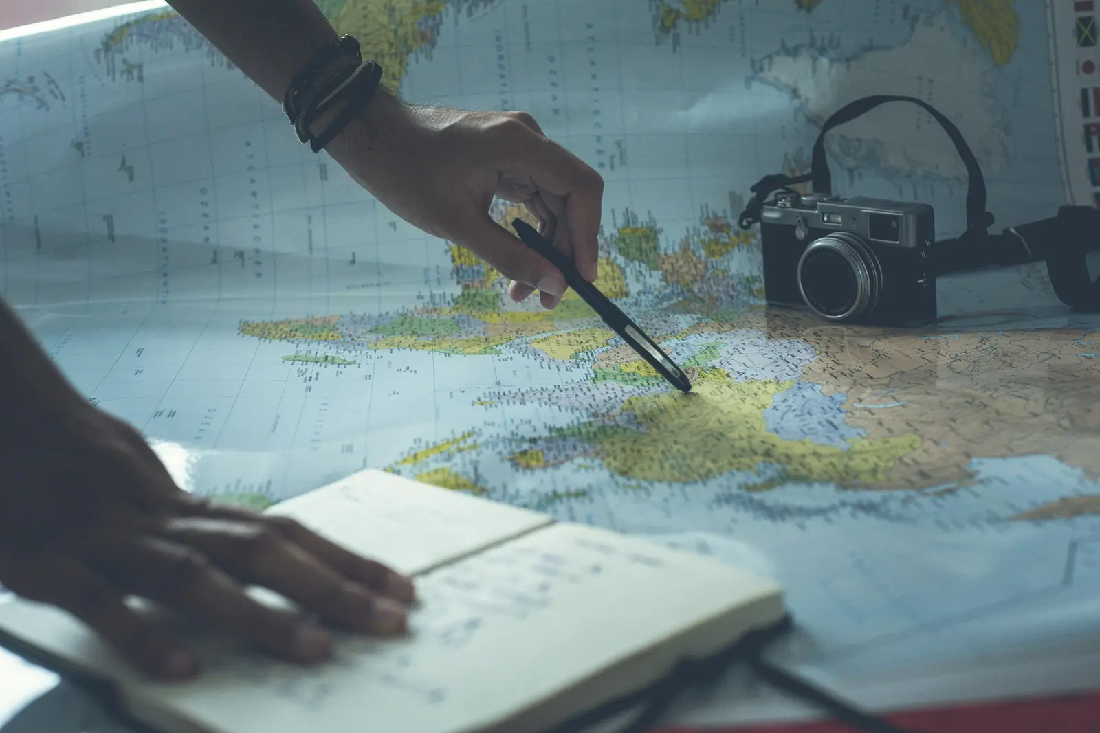 Hand pointing a pen at a printed map of Europe beside a notebook and a compact camera while planning a trip