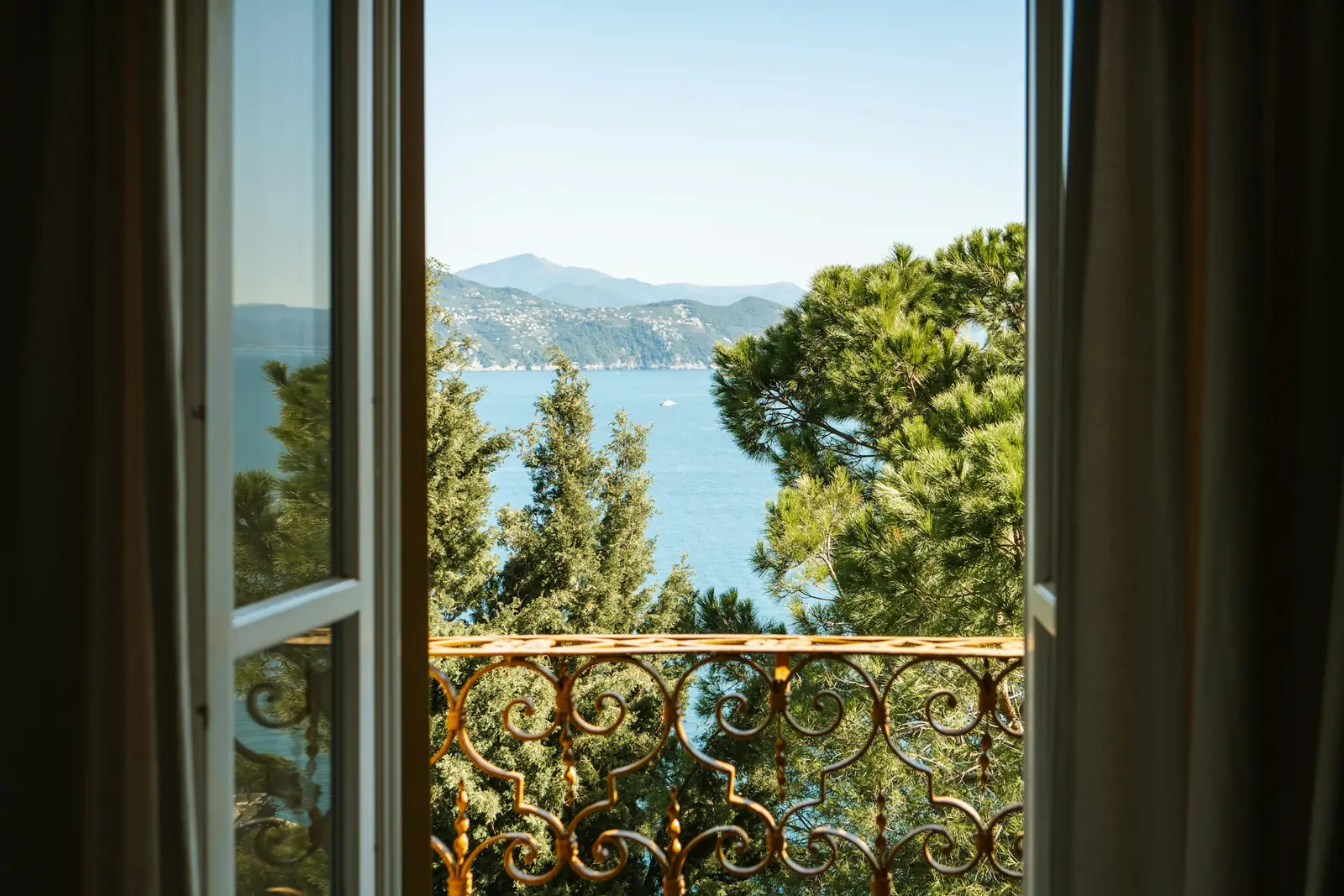 Open French windows onto a wrought-iron balcony overlooking pine trees, the sea and distant hills