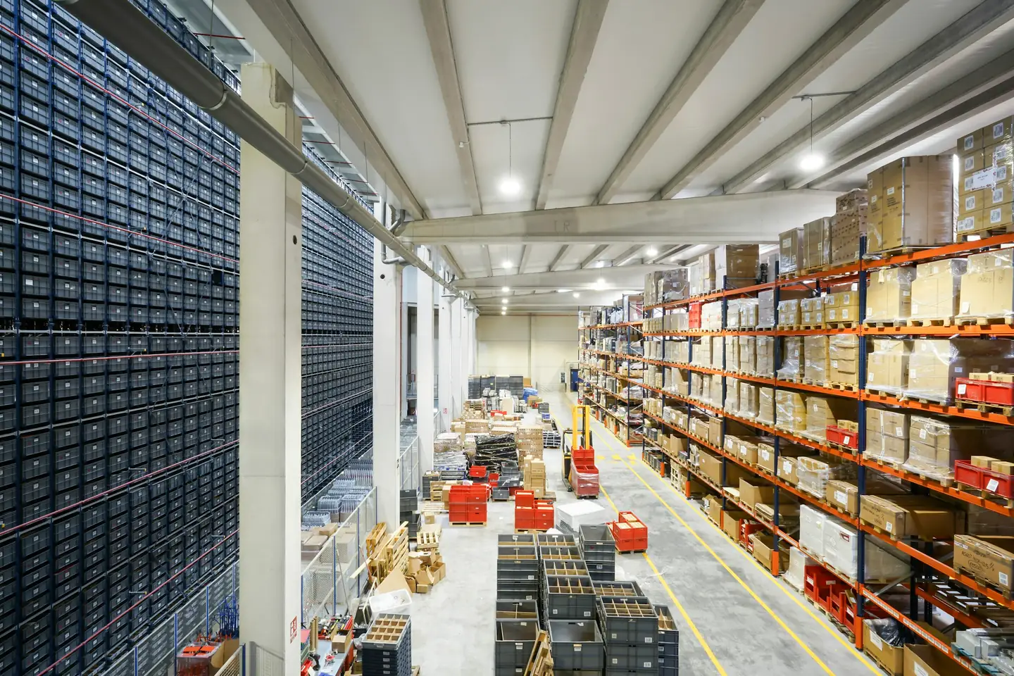 High-bay distribution centre with a wall of grey storage bins beside orange pallet racking and a long picking aisle