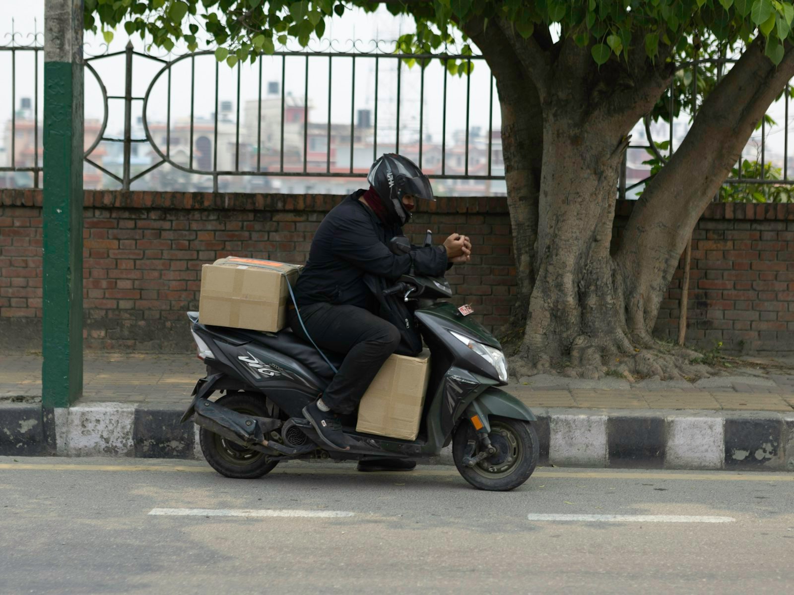 Delivery rider in a helmet on a scooter carrying cardboard parcels along a tree-lined South Asian city street