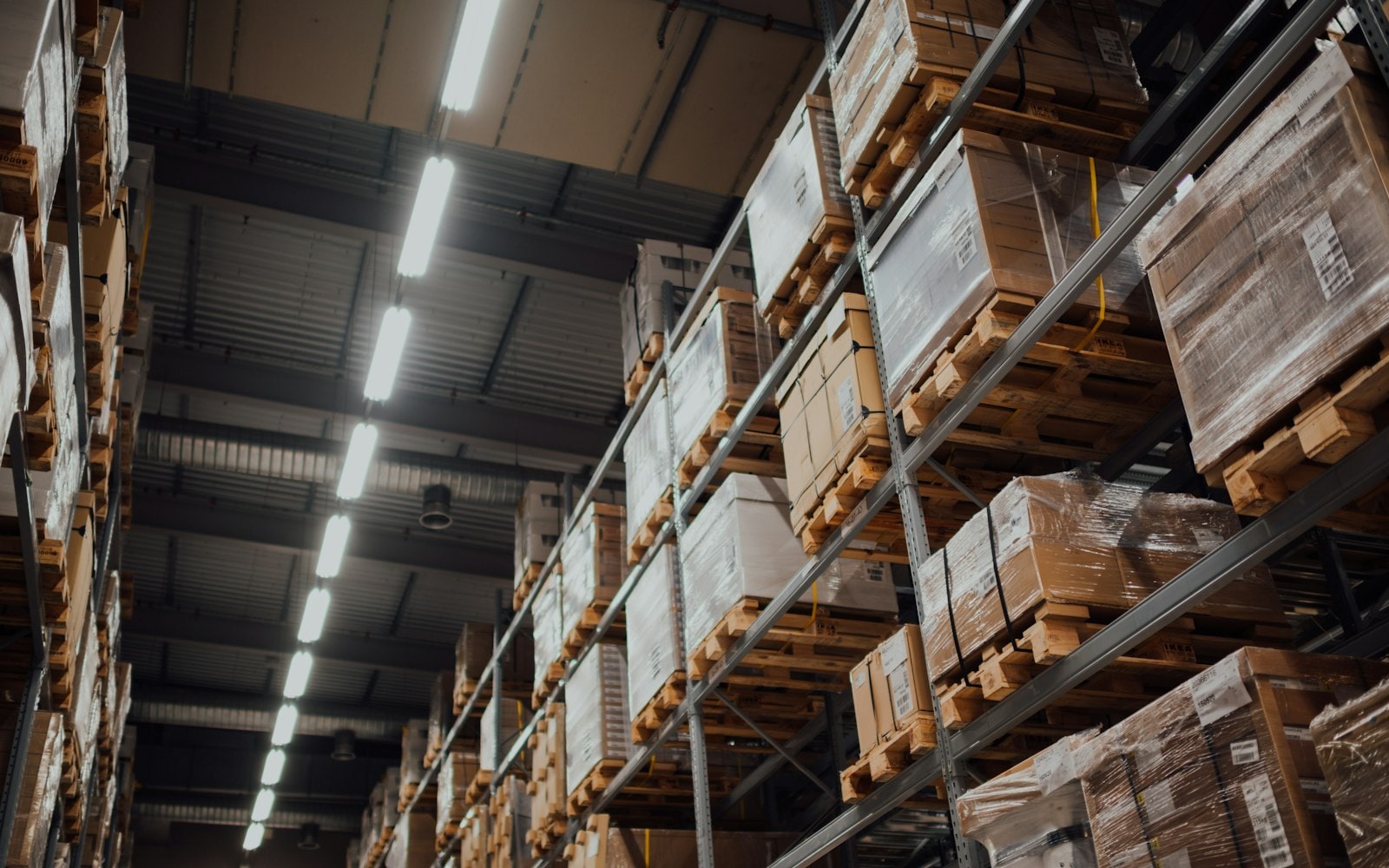 Warehouse racking stacked with shrink-wrapped cartons on wooden pallets under strip lights